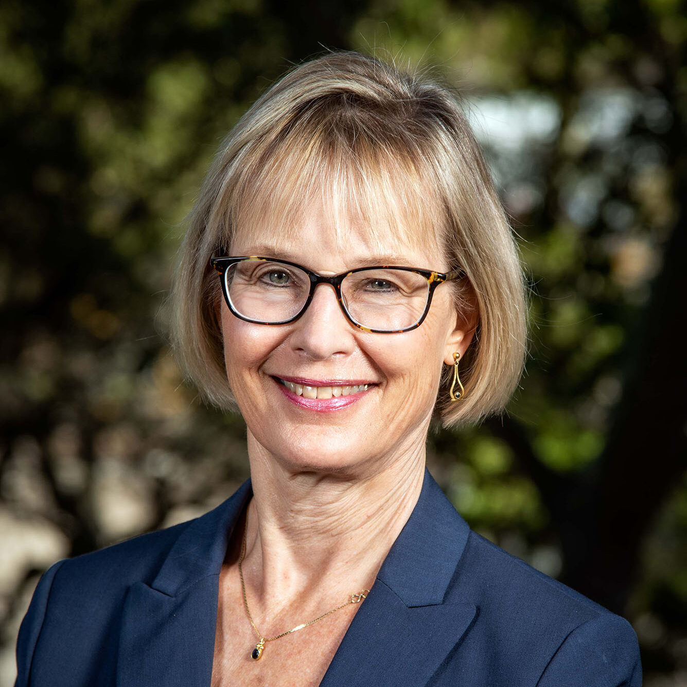 Blonde-haired, blue-eyed person with glasses wearing a navy suit.