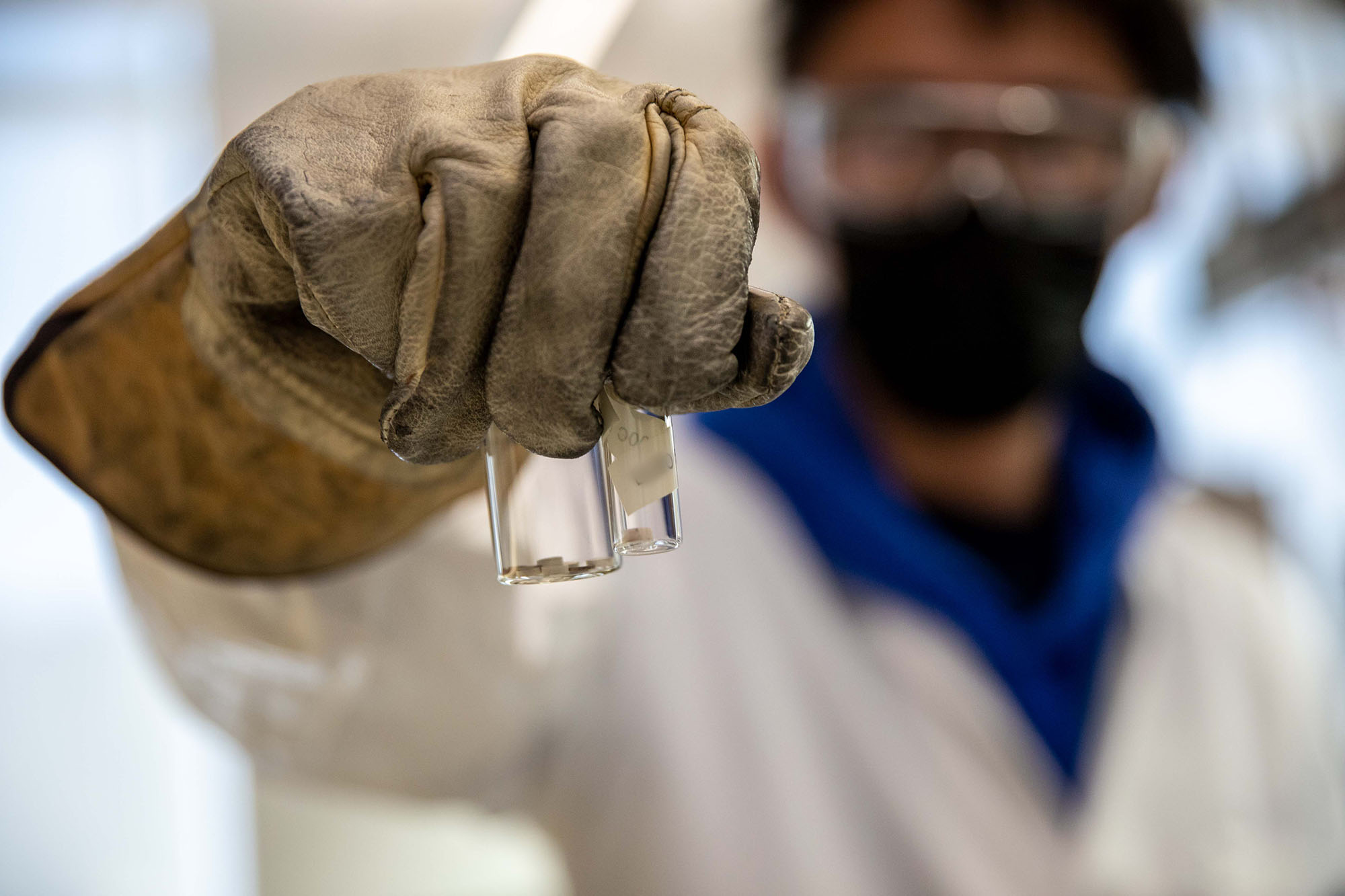 A lab scientist wearing protective equipment holding out two glass viles.