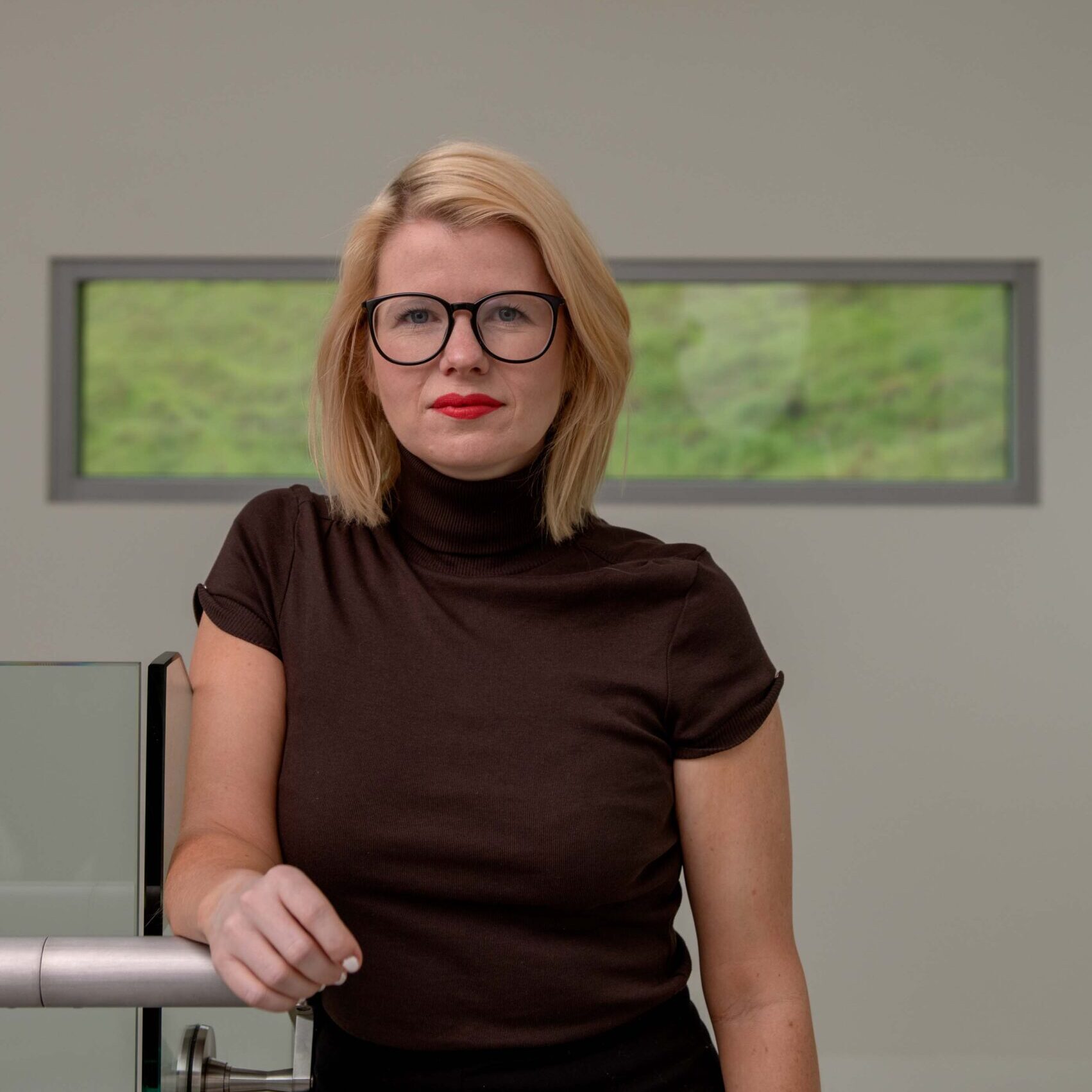 Blonde-haired person in glasses standing in front of a windowed wall.