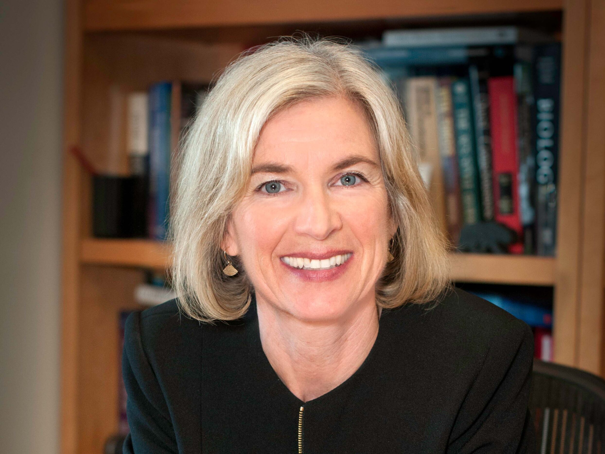 Jennifer Doudna, a light-haired person wearing a black jacket, smiles in front of a bookcase.