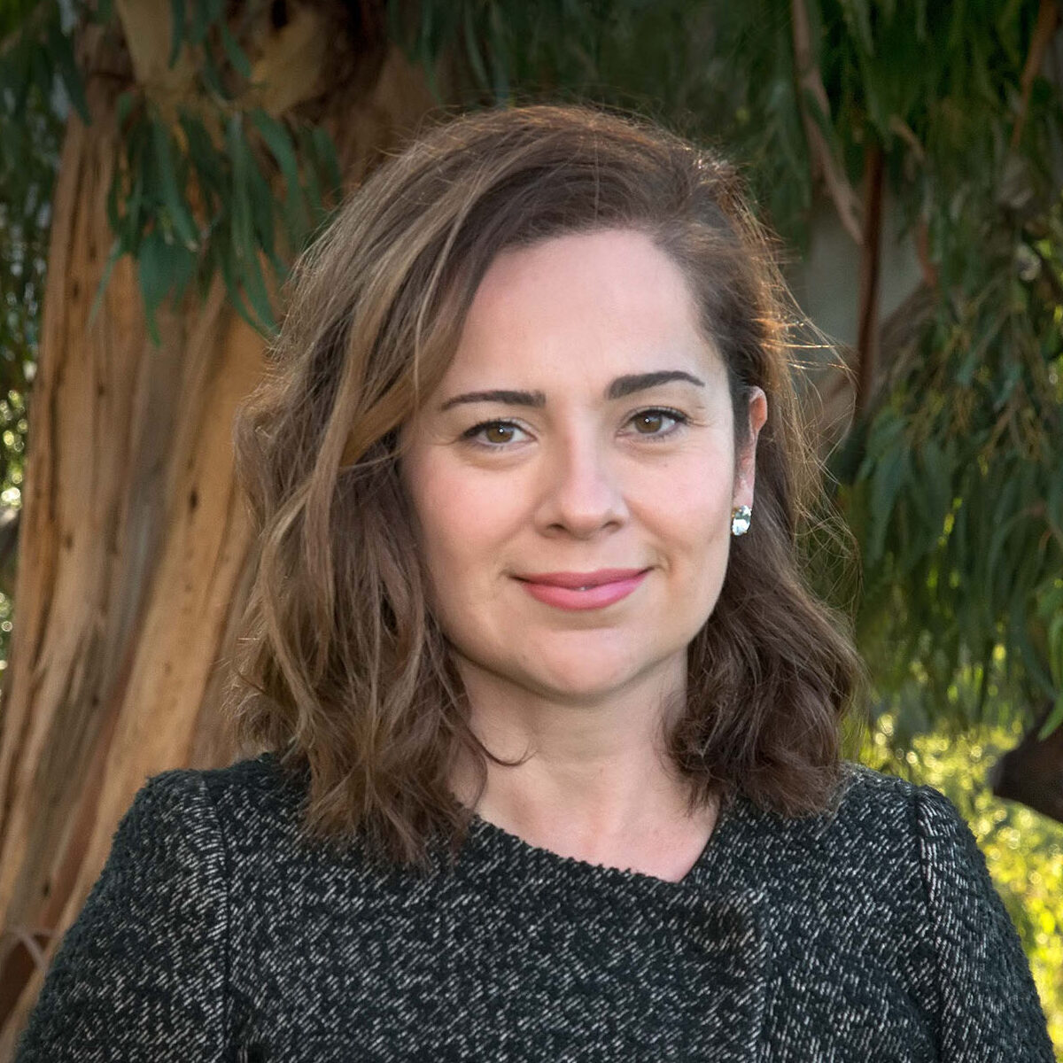 Dark-haired person wearing a black jacket in front of a tree.