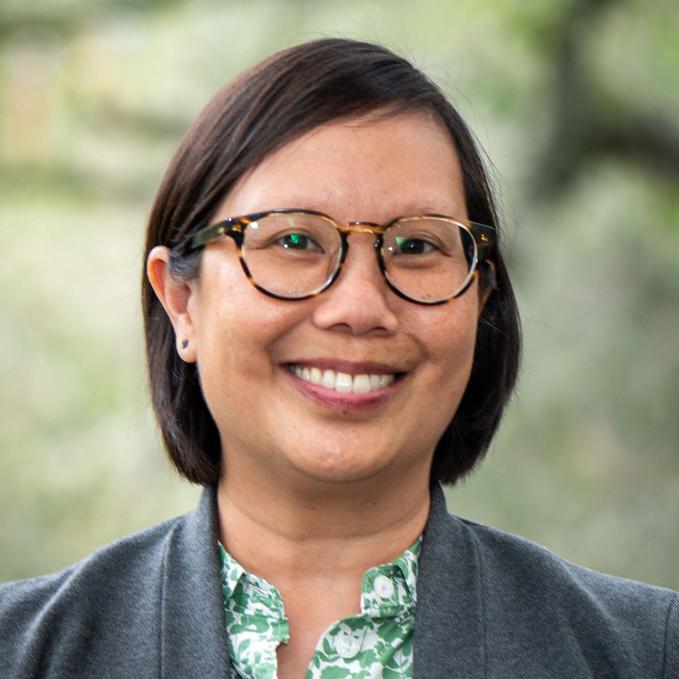 Dark-haired person with glasses wearing green blouse and gray suit.