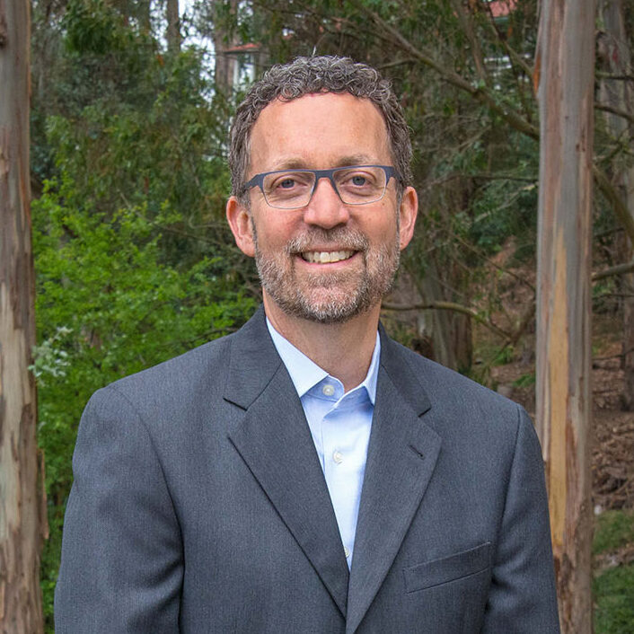 Dark-haired person with glasses wearing a light blue shirt and gray suit jacket.