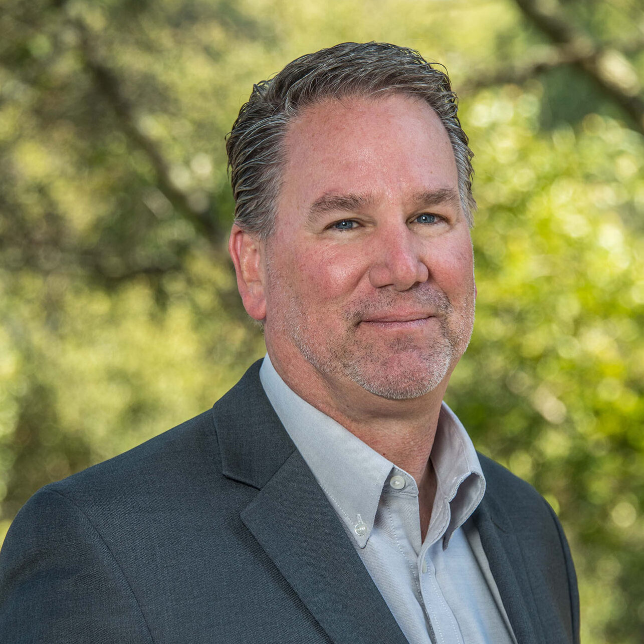 Person wearing a dark gray suit in front of green foliage.