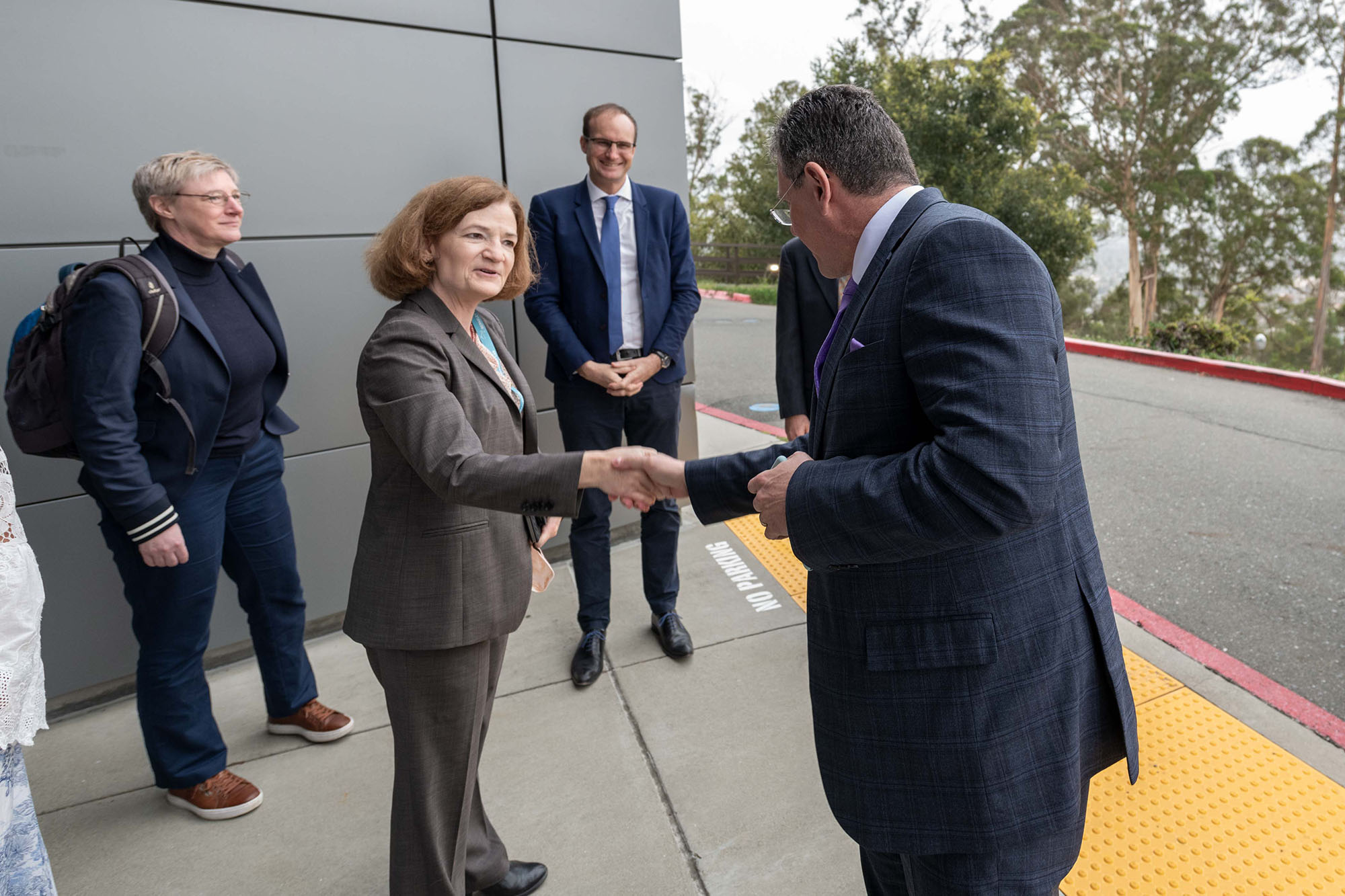 Person with auburn hair in a gray suit shakes hands with a person in a plaid, navy suit in front of two onlookers.