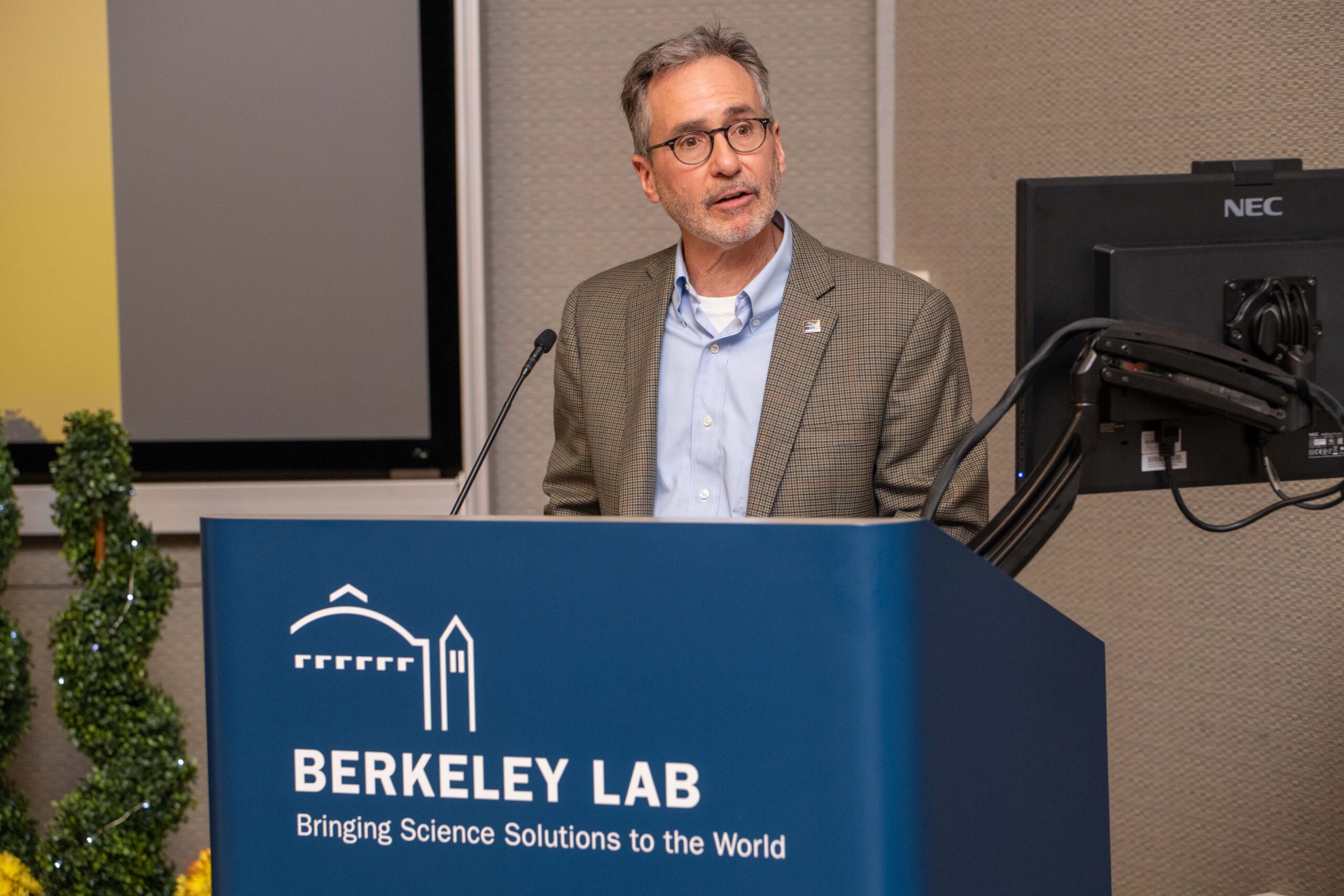 Person in a gray suit, light blue shirt, and glasses stands at a podium and reads from a computer monitor.