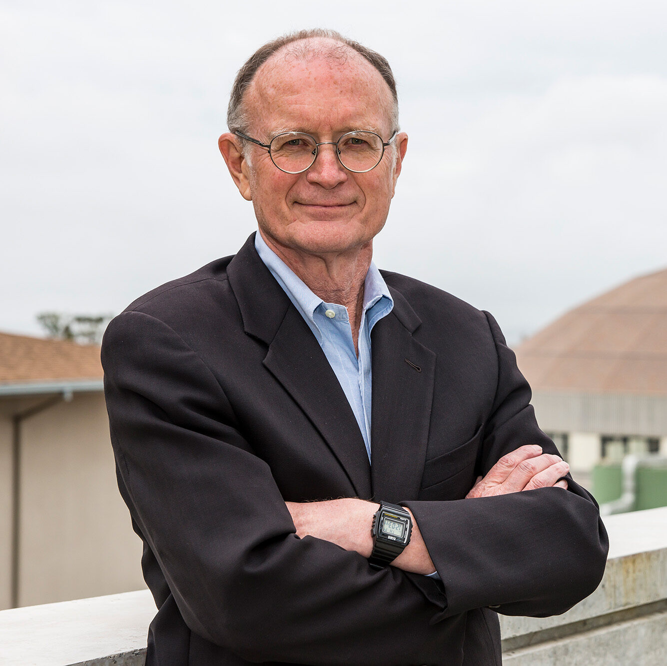 Person in a black suit and glasses standing cross-armed in front of buildings.