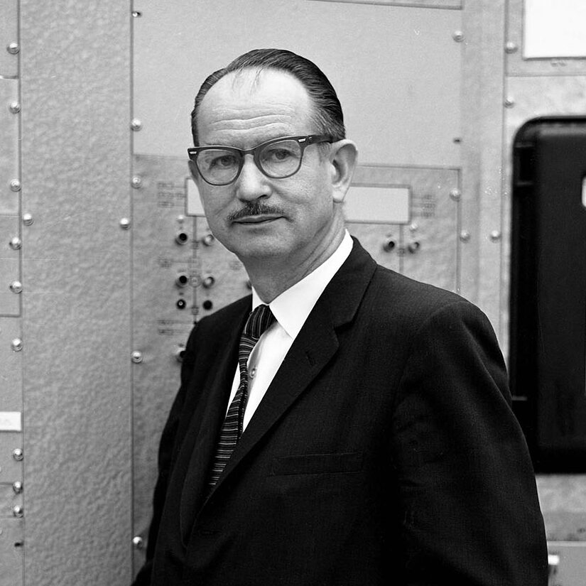 Short-haired person with glasses in a black suit next to lab equipment.