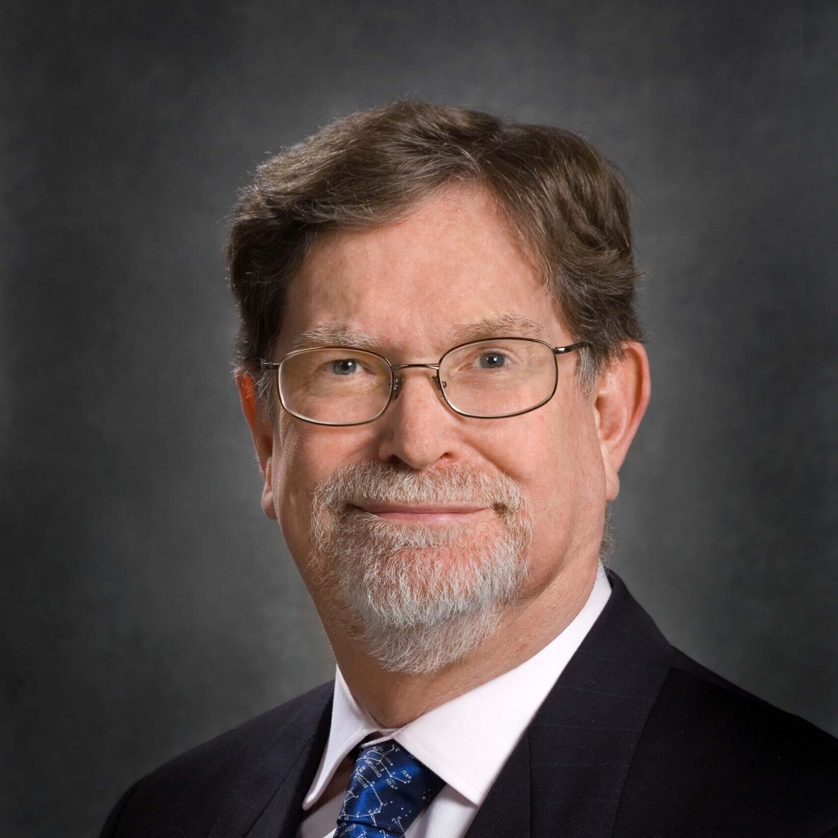 Dark-haired person with glasses wearing a black suit and navy tie.