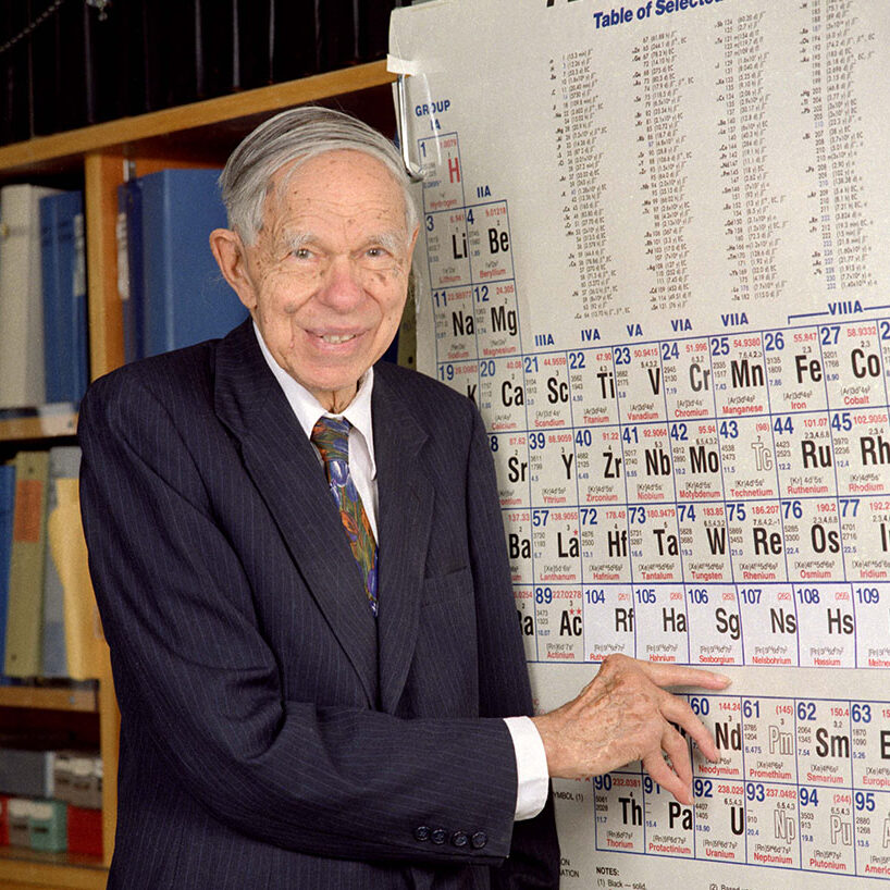 Glenn T. Seaborg, a short-haired person wearing a suit and tie, points to Seaborgium on a period table.