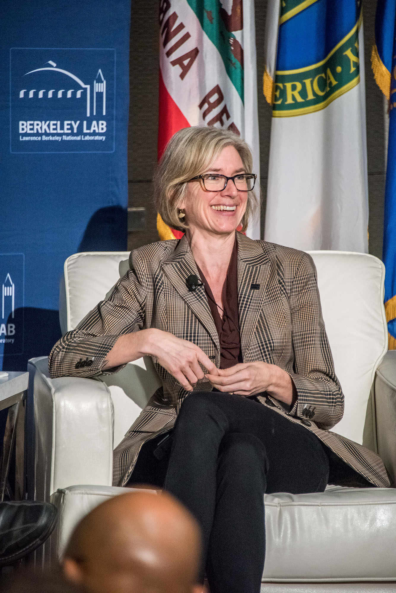 Blonde-haired person in a plaid suit sitting on a white armchair at a press event.