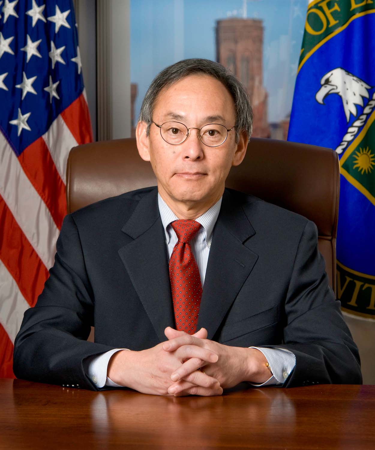 Dark-haired person in a navy suit and red tie sitting in a leather chair.