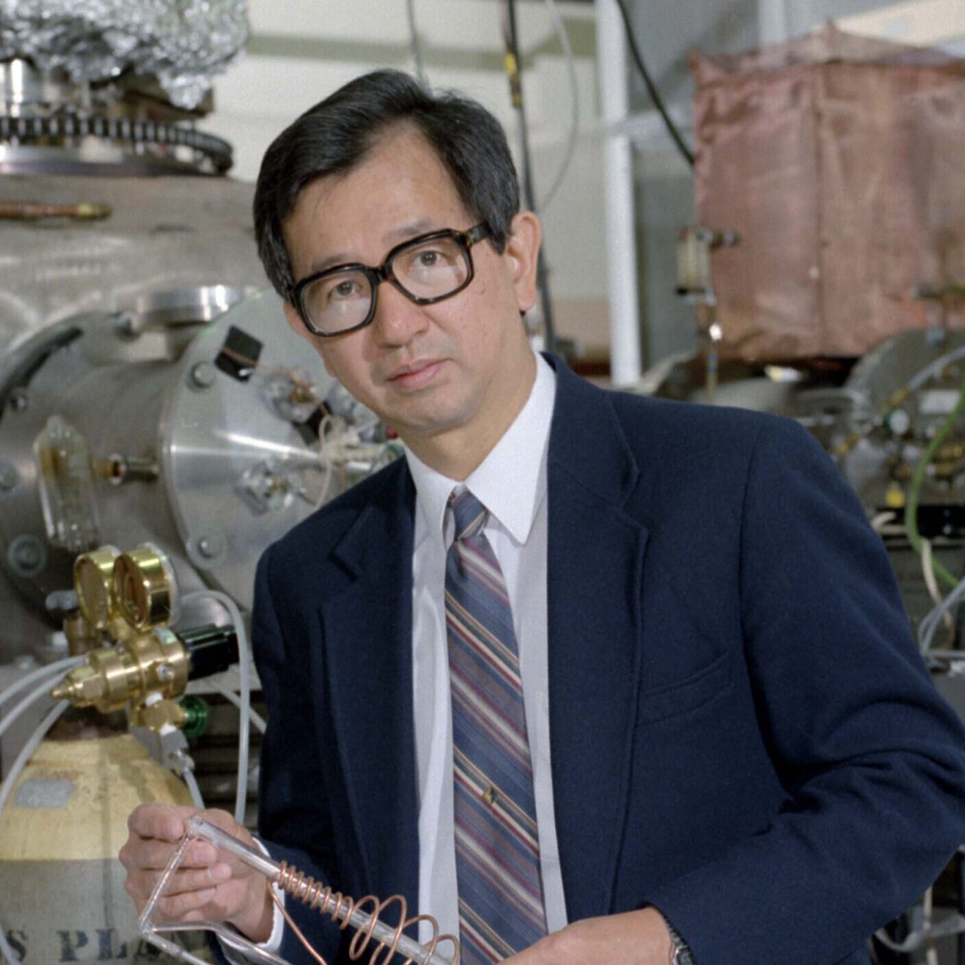 Dark-haired person in glasses standing in a room with machinery.