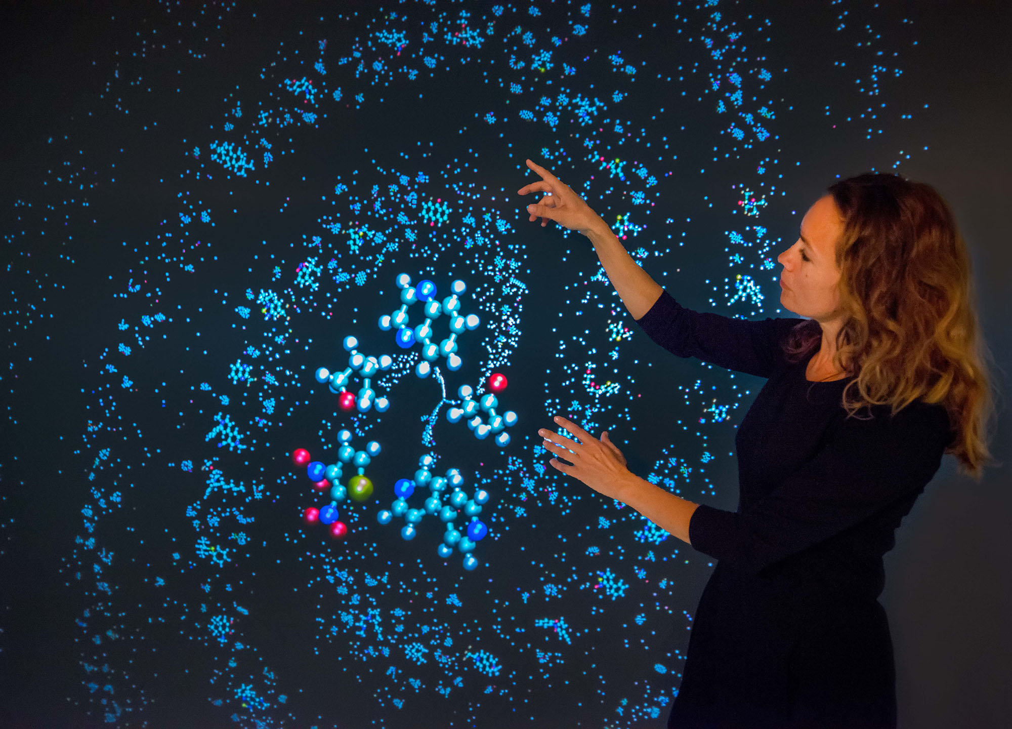 Kristin Persson, a brown-haired person wearing a black dress, points at her electrolyte genome 3D visualizations.
