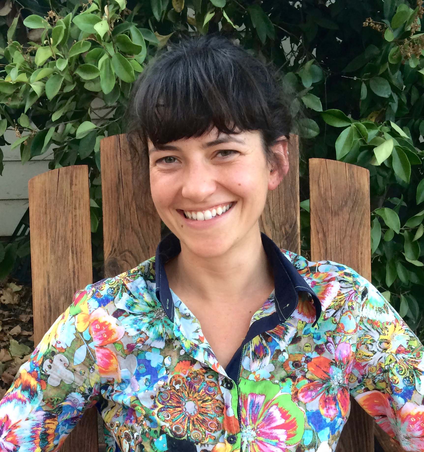 Natalie Popovich, a dark-haired person wearing a colorful button up shirt, smiles against a tree.