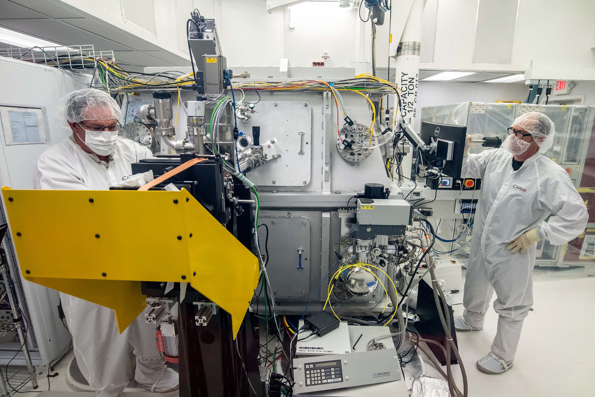 Two researchers in clean suits stand on opposite ends of a large machine.