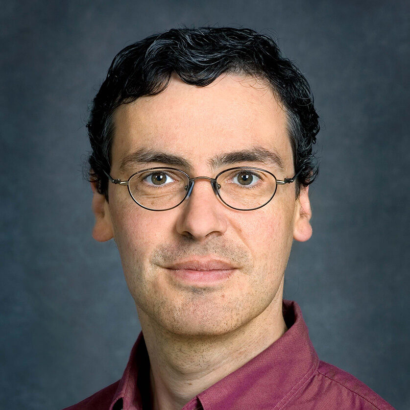 Patrick Naulleau, a short-haired person wearing a red button up shirt against a gray background.