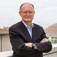 Person in a black suit and glasses standing cross-armed in front of buildings.