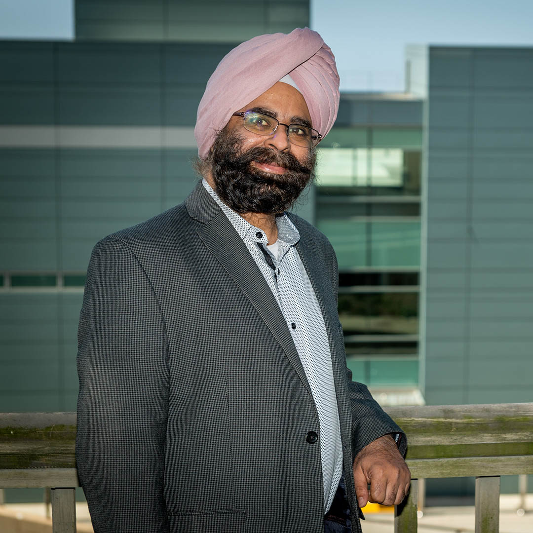 Person with dark hair and a pink turban stands in front of a blue building.