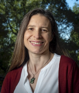 Susannah Tringe, a long brown-haired person wearing a white blouse and red cardigan, smiles outdoors.