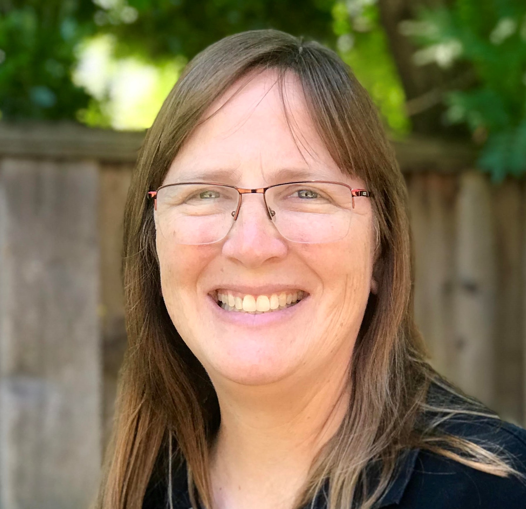 Ann Almgren, a brown-haired person wearing a dark shirt, smiles for a headshot outdoors.