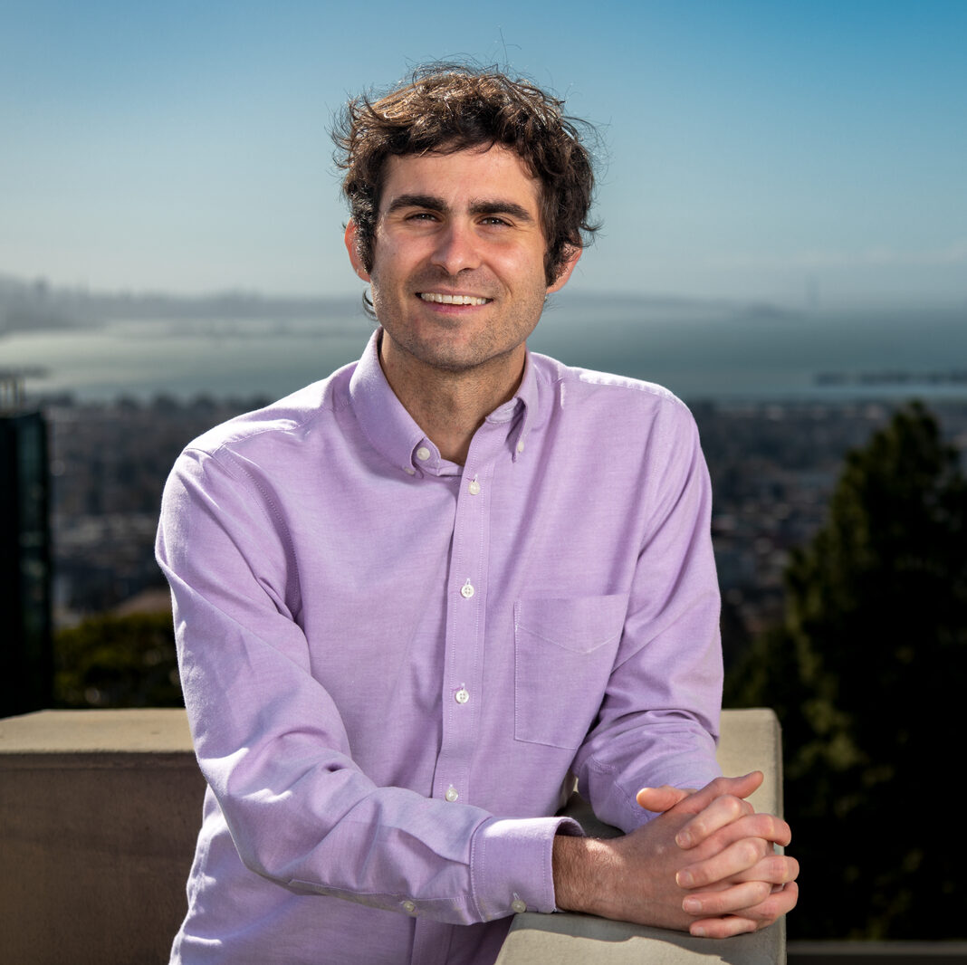 Andrew Haddad, a dark-haired person wearing a purple collared shirt, smiles with the Bay in the background.