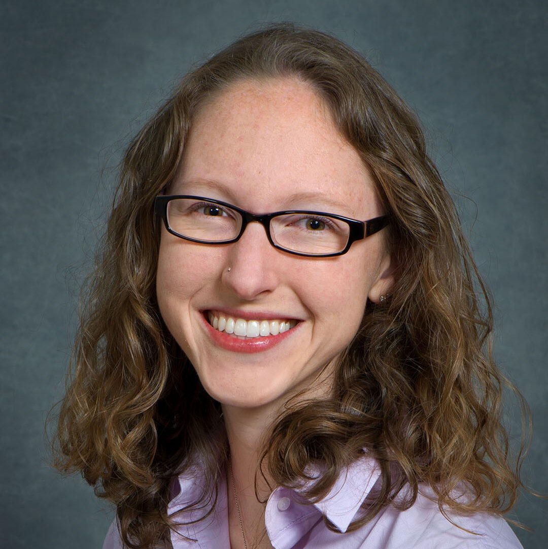 Corinne Scown, a brown-haired person wearing glasses and a purple collared shirt, smiles for a headshot in front of a gray background.