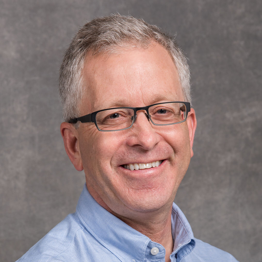 Henrik von der Lippe, a light-haired person wearing dark glasses and a blue collared shirt, smiles for a headshot in front of a gray background.