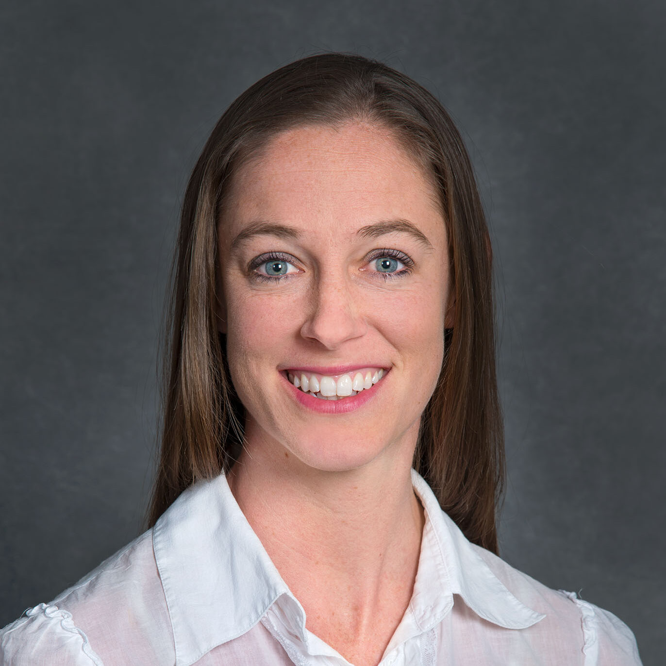 Jennifer Holm, a brown-haired person wearing a white collared shirt, smiles for a headshot against a gray background.