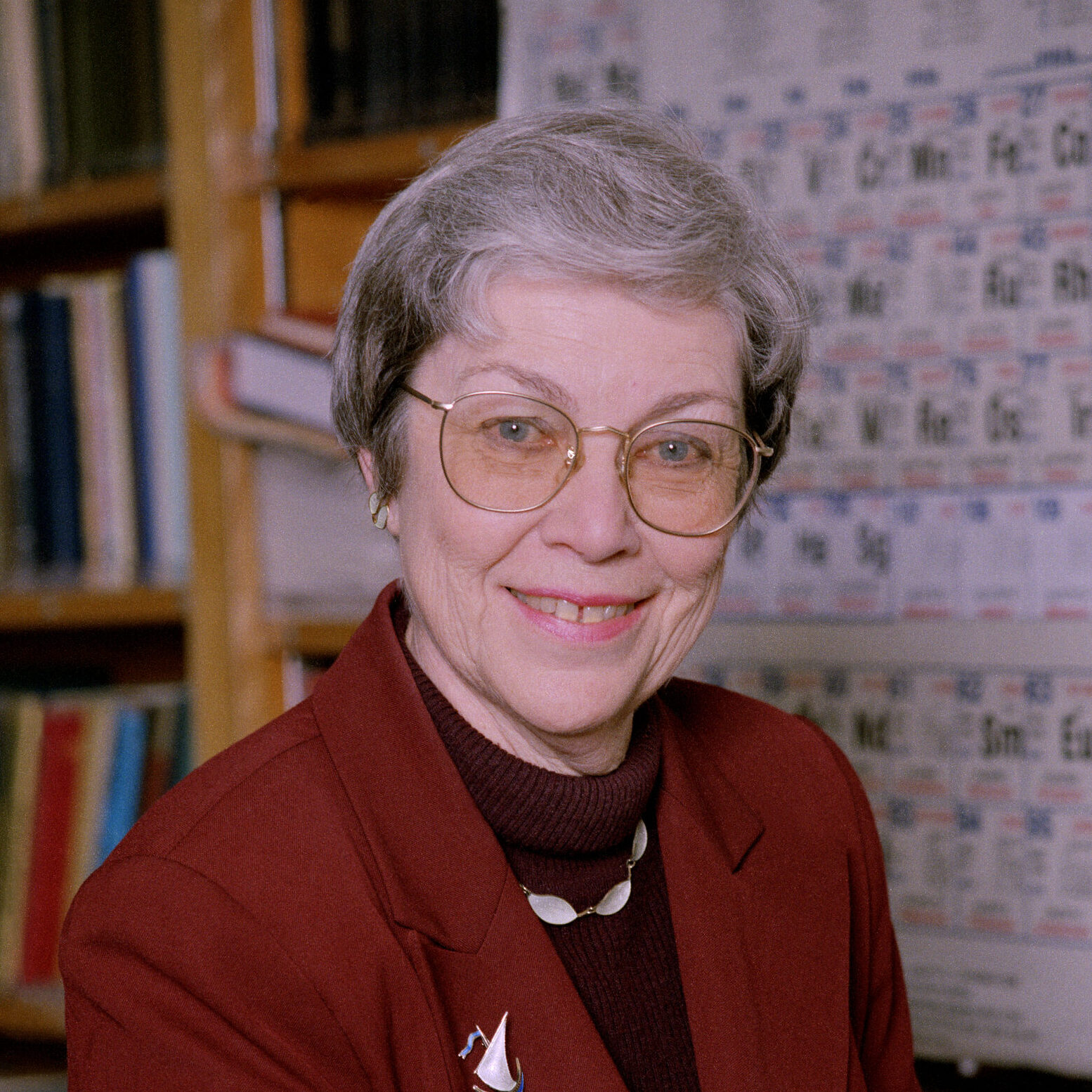 Darleane Hoffman, a light-haired person wearing a red suit, smiles next to a periodic table chart.