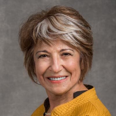 Mina Bissell, light-haired woman wearing a yellow jacket, smiles for her headshot.