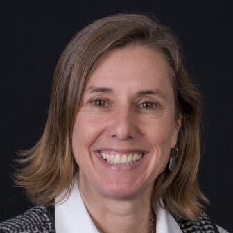 Mary Anne Piette, a brown-haired person wearing a white collared shirt, smiles for a headshot against a dark background.