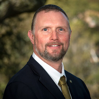 Cameron Geddes, a dark-haired person wearing a black suit, poses for a headshot.