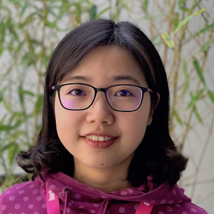Dan Wang, a dark-haired person wearing black glasses and a pink sweater, poses for a headshot in front of a plant.