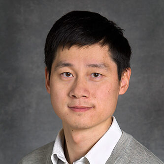 John Wu, a dark-haired person wearing a gray suit, poses for a headshot against a gray background.