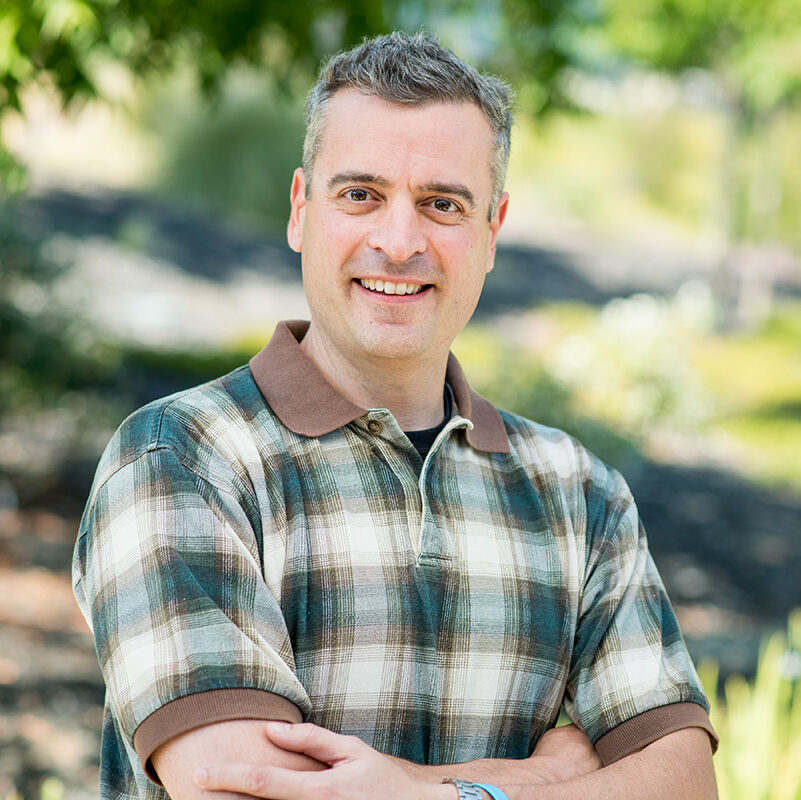 Nathan Hillson, a light-haired person wearing a plaid shirt, poses for a headshot outdoors.
