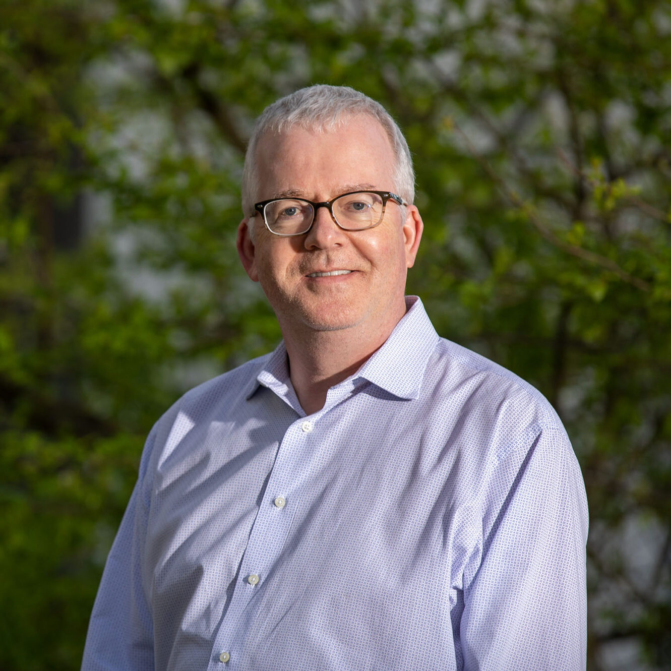 Jonathan Carter, a light haired person wearing a blue collared shirt, poses for a headshot outdoors.