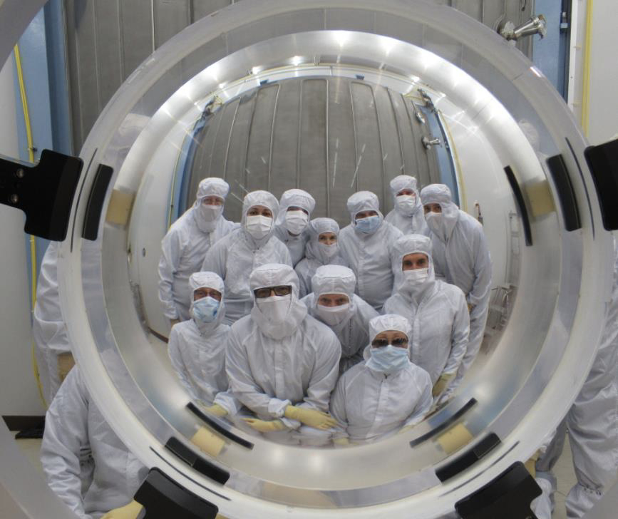 Group of scientists pose behind a large telescope mirror.