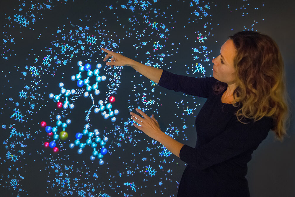 Kristin Persson, a brown-haired person wearing a black dress, points at her electrolyte genome 3D visualizations.