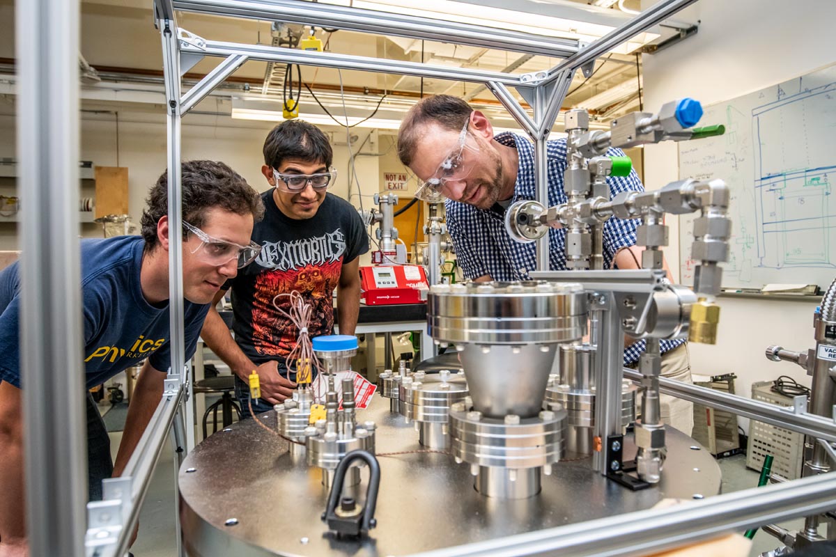 Three scientists standing in a semi-circle around a table of metal instrumentation.