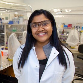 Aindrila Mukhopadhyay, a dark-haired person wearing a white jacket, poses for a headshot in the lab.