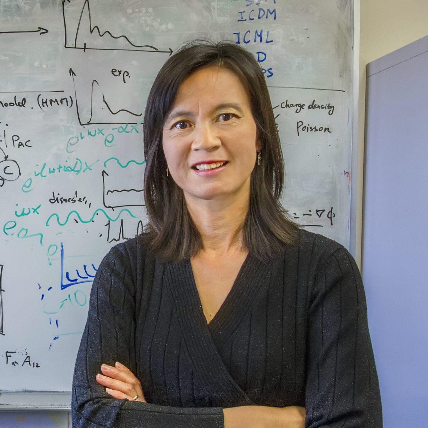 Sherry Li, a dark haired person wearing a black sweater, poses for a headshot in front of a whiteboard.