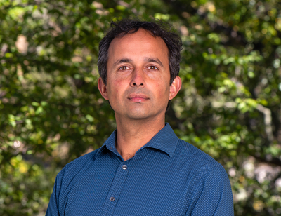 Arun Persaud, a dark-haired person wearing a blue collared shirt, poses for a headshot outdoors.