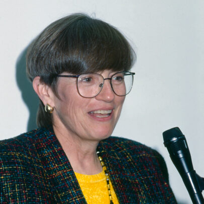 Dark haired person speaking into a microphone.