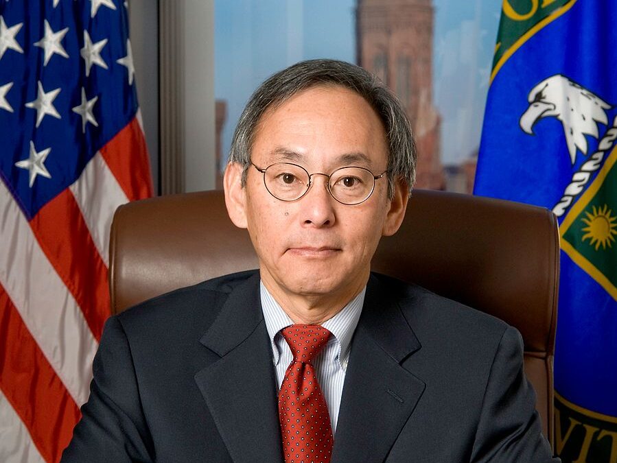 dark-haired person in a black suit and red tie sitting in a leather chair.