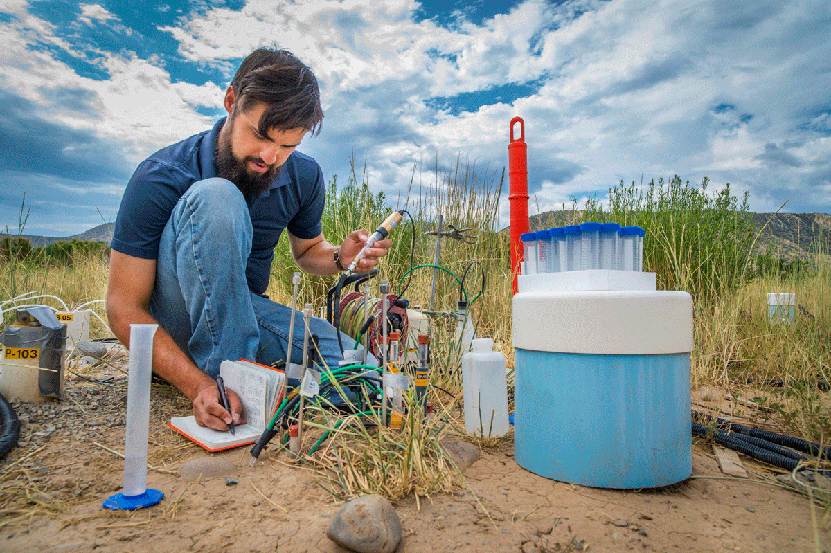 Dark-haired scientist in a polo and jeans kneeling in a field with various instrumentation