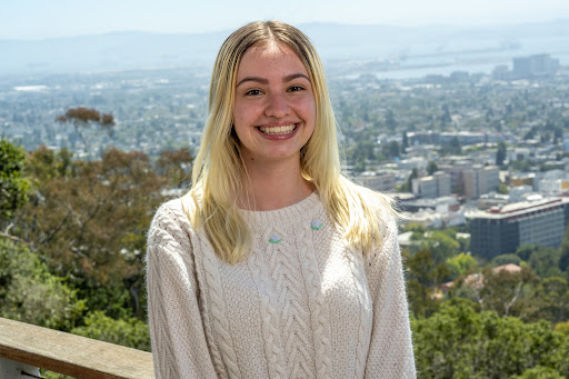 Blonde-haired person in a beige sweater standing in front of a city-scape.