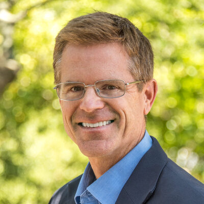 Short-haired person in glasses wearing a blue shirt and black suit.