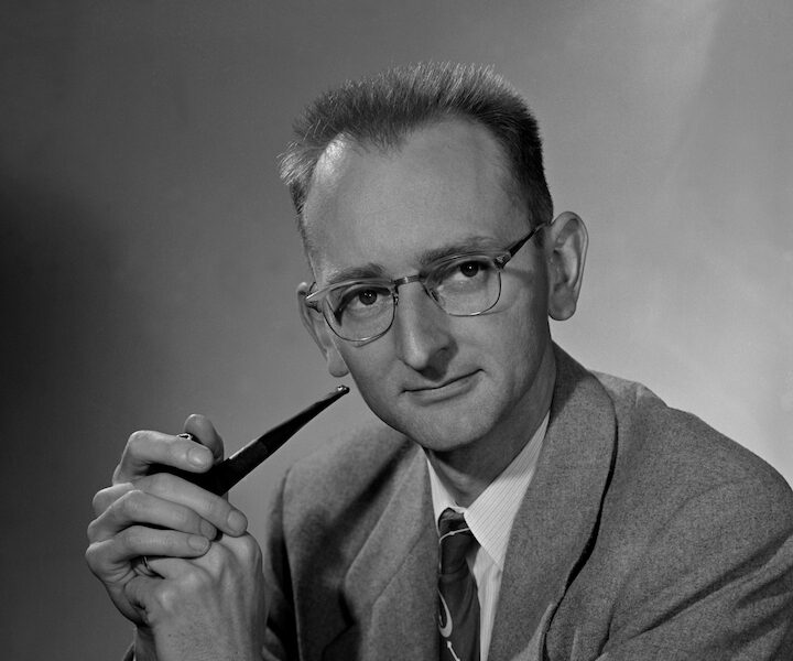 Short-haired person with glasses wearing a suit and holding a pipe.
