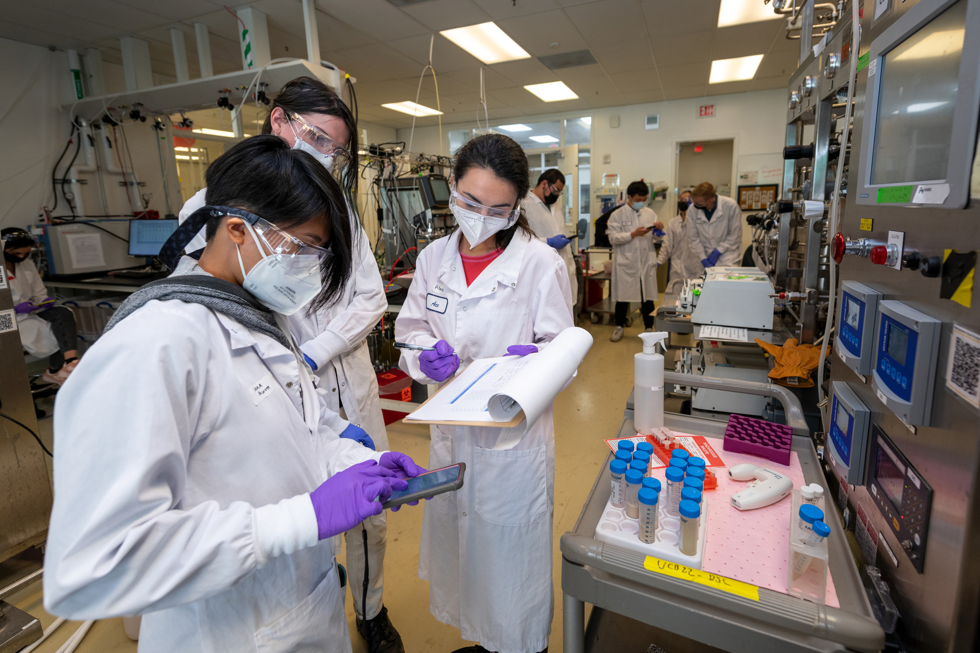 Three students in the foreground look at notes on a clipboard. The group is standing in a laboratory with other students in the background.